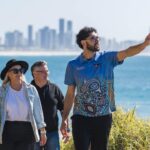 Three people walk outdoors near the coast with a city skyline in the background; the man in front gestures toward the distance.