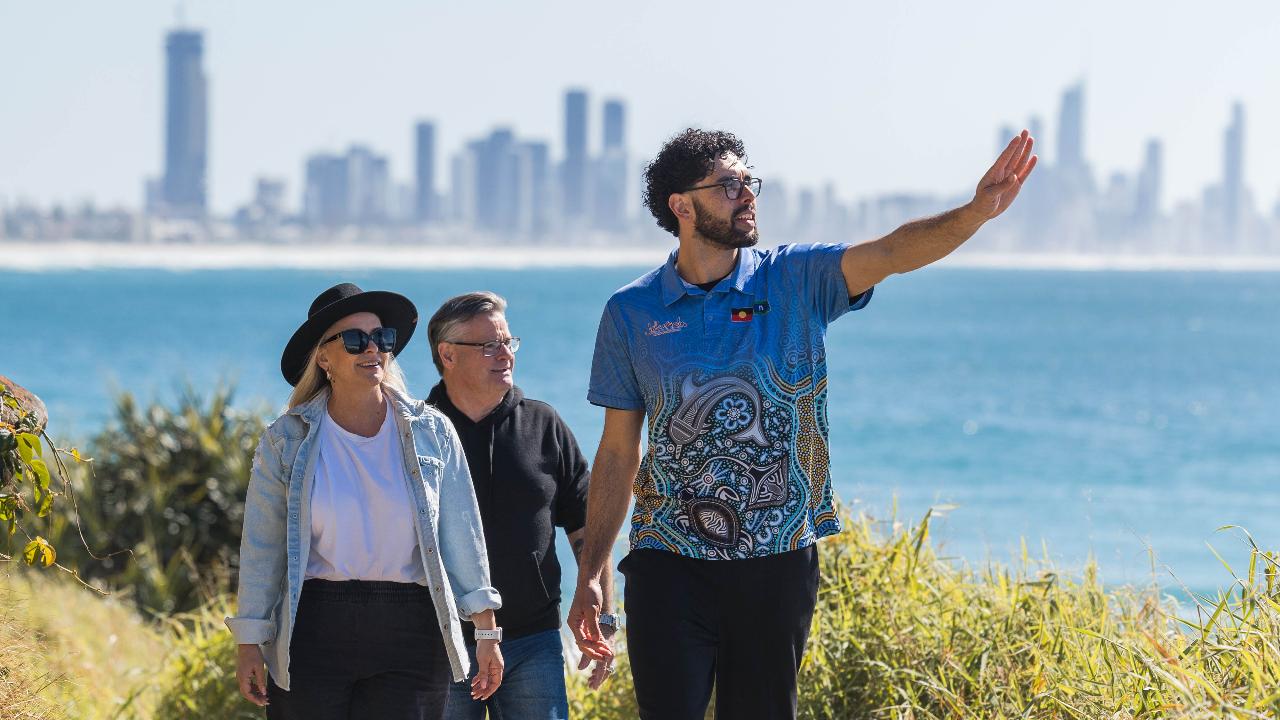 Three people walk outdoors near the coast with a city skyline in the background; the man in front gestures toward the distance.