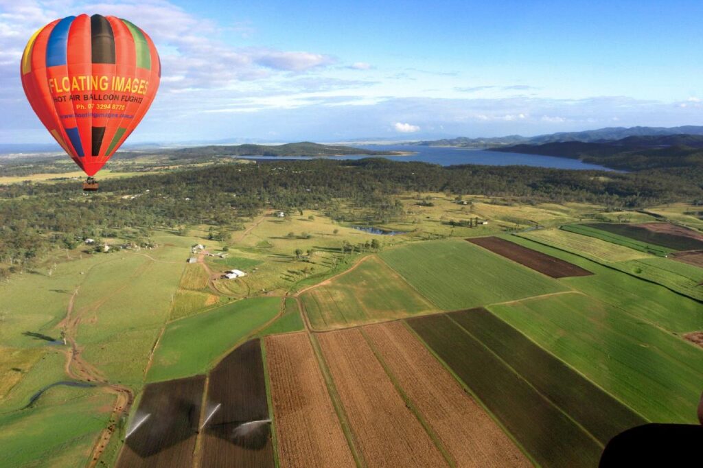 A hot air balloon floats above patchwork farmland with fields, scattered houses, and a body of water visible in the background.