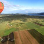 A hot air balloon floats above patchwork farmland with fields, scattered houses, and a body of water visible in the background.