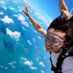 A person wearing goggles and a harness is skydiving over a blue ocean with scattered clouds below.