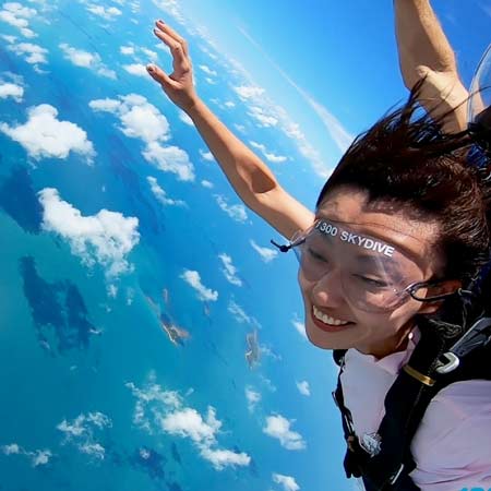 A person wearing goggles and a harness is skydiving over a blue ocean with scattered clouds below.