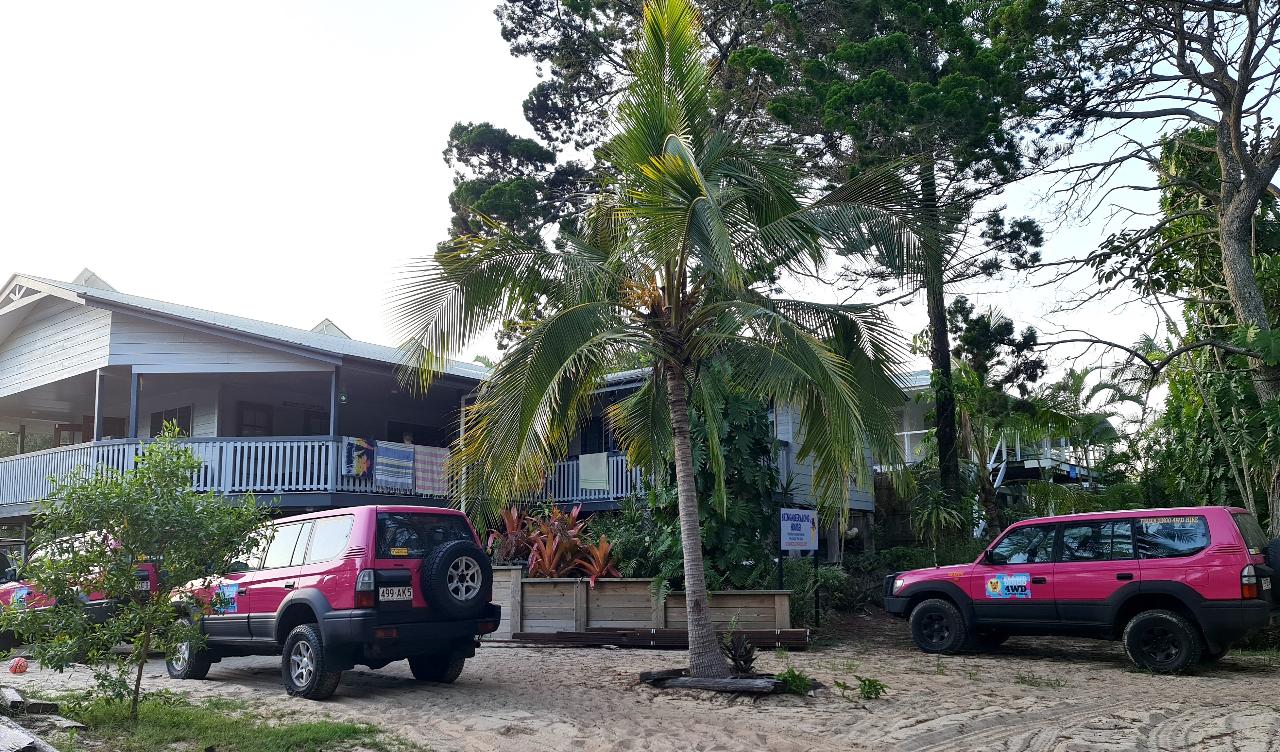 Two pink four-wheel-drive vehicles are parked in front of a tropical-style building surrounded by palm trees and greenery.