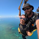 Two people tandem skydiving over an ocean, smiling at the camera with blue sky and water visible below.