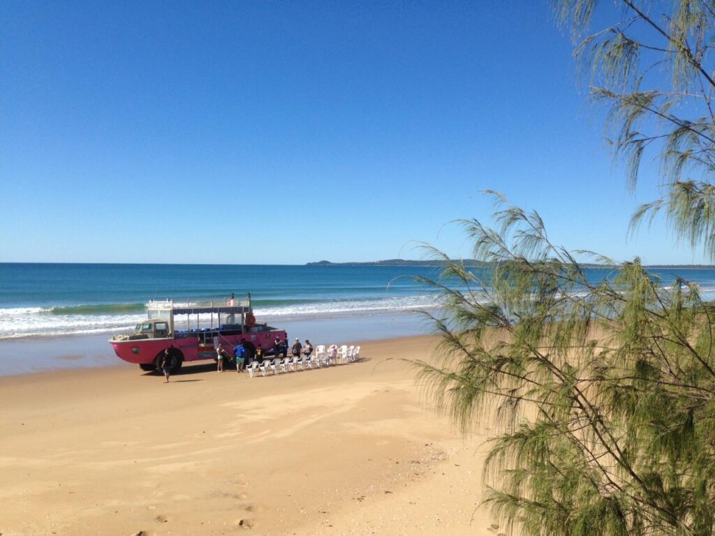 A red amphibious vehicle is parked on a sandy beach near the shore, with chairs and people gathered nearby under a clear blue sky.
