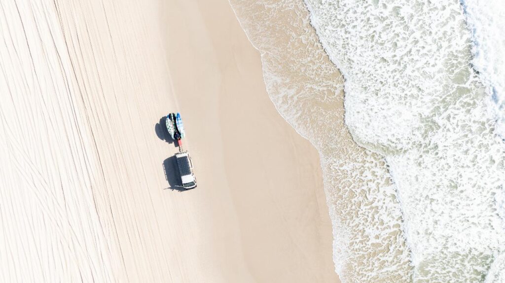 Aerial view of a white vehicle parked on a sandy beach near the shoreline, with tire tracks visible and waves approaching from the right.