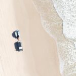 Aerial view of a white vehicle parked on a sandy beach near the shoreline, with tire tracks visible and waves approaching from the right.