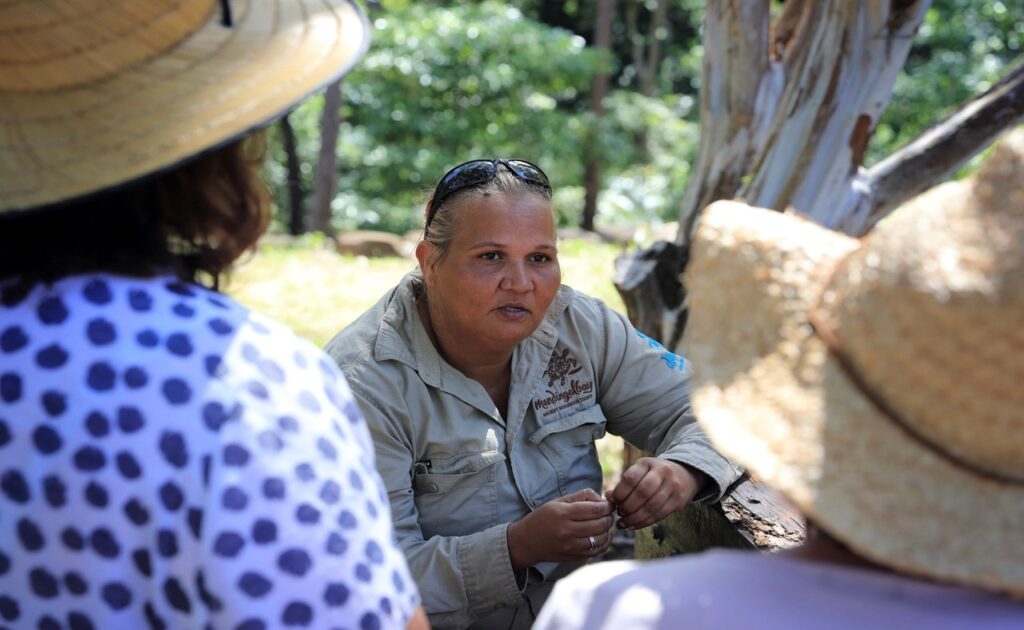 A woman in a beige shirt and sunglasses talks to two people wearing straw hats in an outdoor setting with trees in the background.