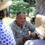 A woman in a beige shirt and sunglasses talks to two people wearing straw hats in an outdoor setting with trees in the background.