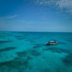 A small boat with passengers floats on clear blue ocean water under a partly cloudy sky, with reef patterns visible beneath the surface.