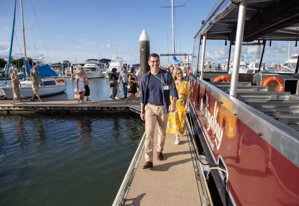 People walk along a dock next to a red boat at a marina, with various other boats and yachts visible in the background under a blue sky.