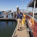 People walk along a dock next to a red boat at a marina, with various other boats and yachts visible in the background under a blue sky.