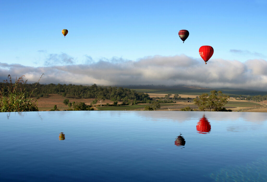 Three hot air balloons float above a rural landscape under a blue sky, with their reflections visible in the still water of a pool in the foreground.