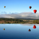 Three hot air balloons float above a rural landscape under a blue sky, with their reflections visible in the still water of a pool in the foreground.
