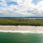 Aerial view of a sandy beach with waves, bordered by a strip of trees and dense greenery under a partly cloudy sky.