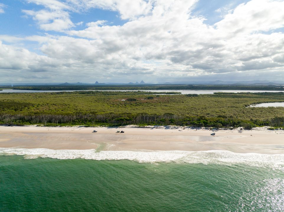 Aerial view of a sandy beach with waves, bordered by a strip of trees and dense greenery under a partly cloudy sky.