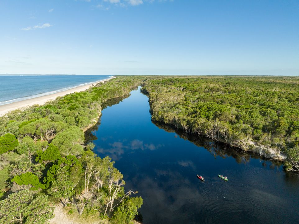 Aerial view of a river flowing through dense green forest beside a sandy beach, with two kayaks visible on the water under a clear blue sky.