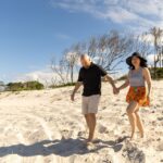 A man and woman walk hand in hand on a sandy beach with sparse trees in the background under a partly cloudy sky.