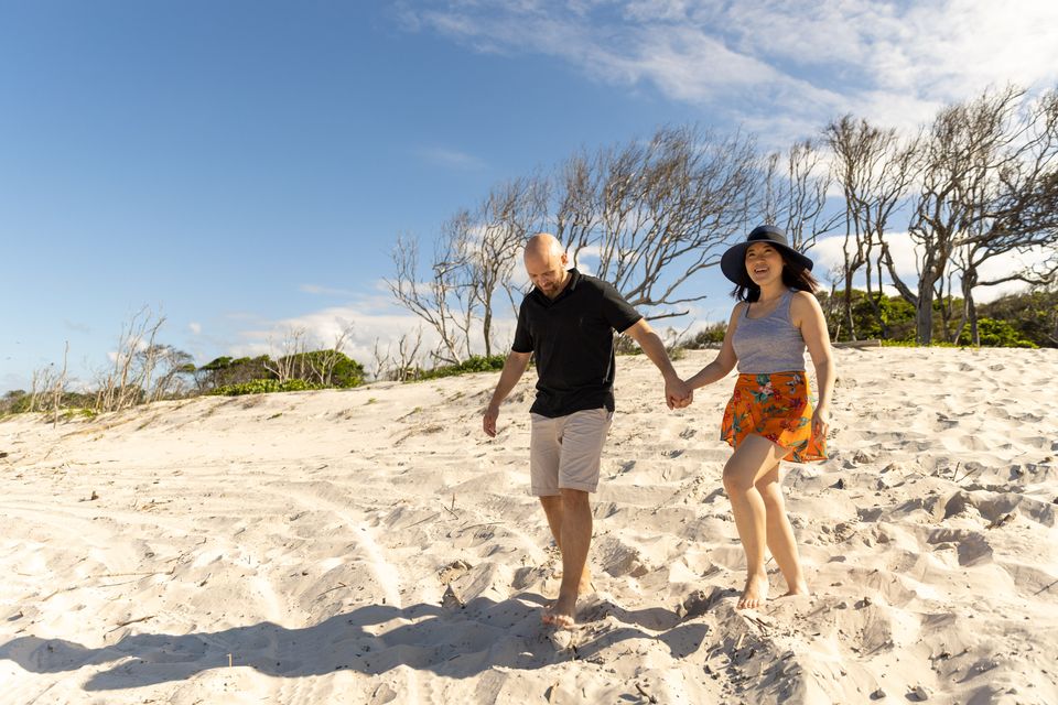 A man and woman walk hand in hand on a sandy beach with sparse trees in the background under a partly cloudy sky.