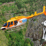A yellow and white Zoom helicopter flies over a lush green landscape with a waterfall in the background.