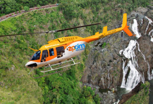 A yellow and white Zoom helicopter flies over a lush green landscape with a waterfall in the background.