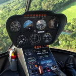 Close-up view of a helicopter cockpit instrument panel with various gauges and controls, flying over a green landscape with trees and an open field visible through the windshield.