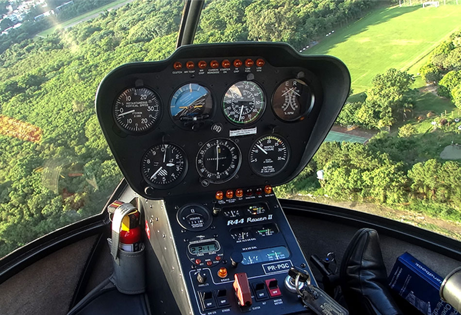 Close-up view of a helicopter cockpit instrument panel with various gauges and controls, flying over a green landscape with trees and an open field visible through the windshield.