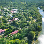 Aerial view of a small town surrounded by dense greenery, with buildings near a winding river and a bridge crossing the water.