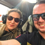 Two people wearing sunglasses and headsets sit inside a helicopter; the woman on the left makes a peace sign while the man on the right smiles at the camera.