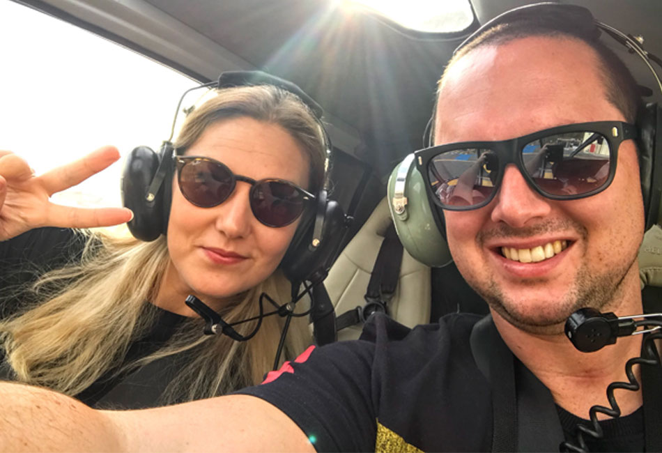 Two people wearing sunglasses and headsets sit inside a helicopter; the woman on the left makes a peace sign while the man on the right smiles at the camera.