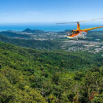 A yellow helicopter flies over a green forested hillside with a coastal town and ocean visible in the background under a clear blue sky.