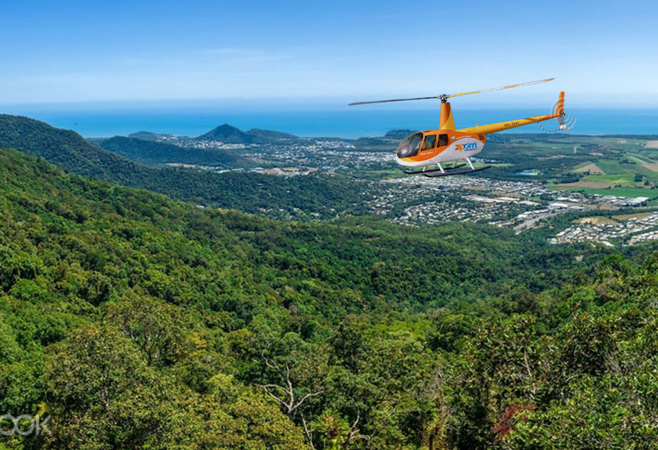A yellow helicopter flies over a green forested hillside with a coastal town and ocean visible in the background under a clear blue sky.