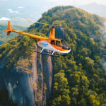An orange and white helicopter flies over a dense forest with rocky cliffs and mountains in the background.
