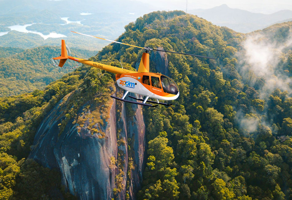 An orange and white helicopter flies over a dense forest with rocky cliffs and mountains in the background.