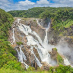 A wide waterfall cascades over rocky cliffs surrounded by dense green forest under a partly cloudy sky.