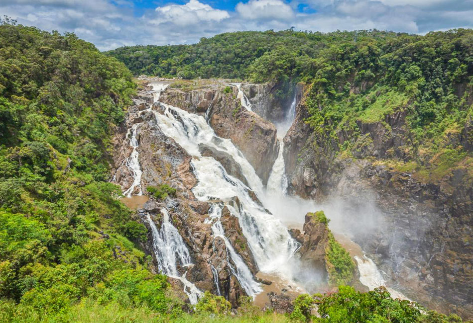A wide waterfall cascades over rocky cliffs surrounded by dense green forest under a partly cloudy sky.
