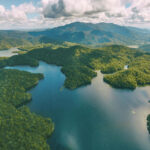 Aerial view of a lake surrounded by dense green forest and mountains under a partly cloudy sky.