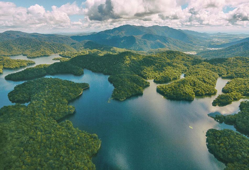 Aerial view of a lake surrounded by dense green forest and mountains under a partly cloudy sky.