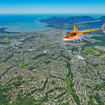 A yellow helicopter flies over a coastal city with houses, roads, and greenery, with the ocean and mountains visible in the background.