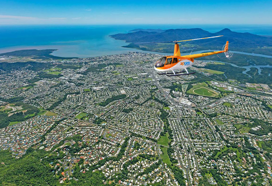 A yellow helicopter flies over a coastal city with houses, roads, and greenery, with the ocean and mountains visible in the background.