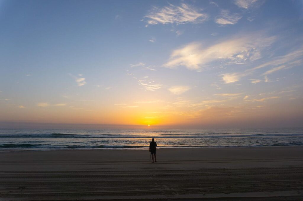 A person stands on a sandy beach facing the ocean at sunrise, with calm waves and a partly cloudy sky.