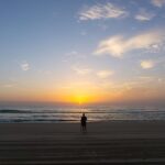 A person stands on a sandy beach facing the ocean at sunrise, with calm waves and a partly cloudy sky.