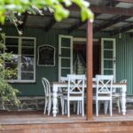 A wooden porch with a white dining table and chairs, green siding, open double doors, and a chalkboard sign on the wall, partially shaded by leafy branches.