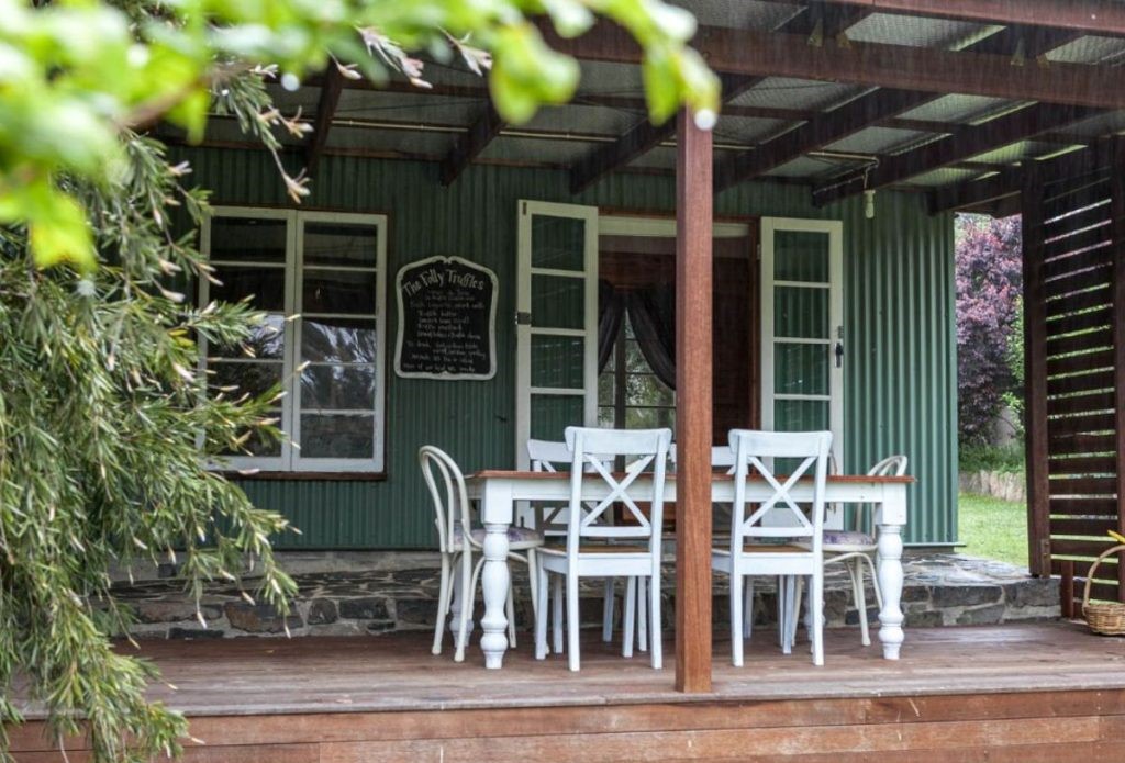 A wooden porch with a white dining table and chairs, green siding, open double doors, and a chalkboard sign on the wall, partially shaded by leafy branches.