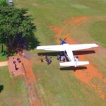 A small airplane is parked on a grassy field as several people walk to and from the aircraft near a fenced area and a tree.