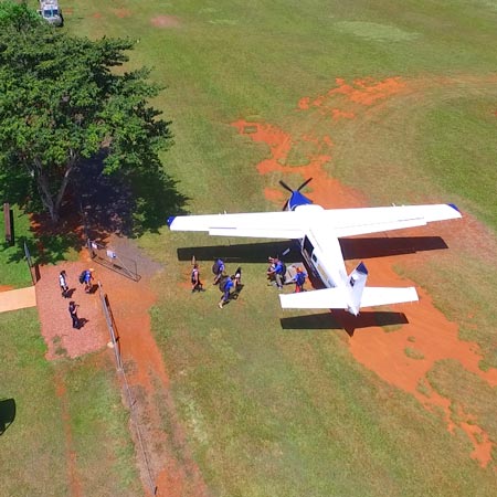 A small airplane is parked on a grassy field as several people walk to and from the aircraft near a fenced area and a tree.