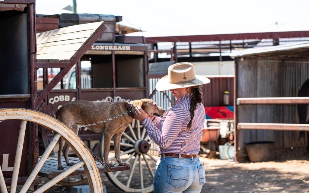 A woman in a hat pets a dog that is standing on the step of a wooden wagon, with farm buildings visible in the background.