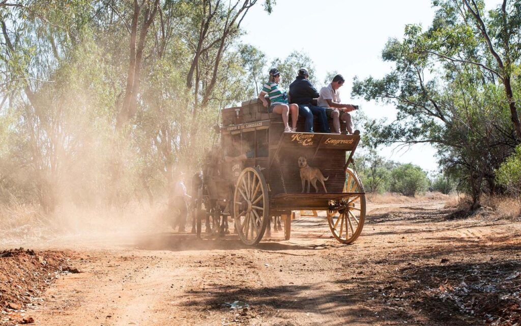 A horse-drawn stagecoach travels on a dusty dirt road with several people sitting on top and a dog standing on the front. Trees and dry vegetation line the path.