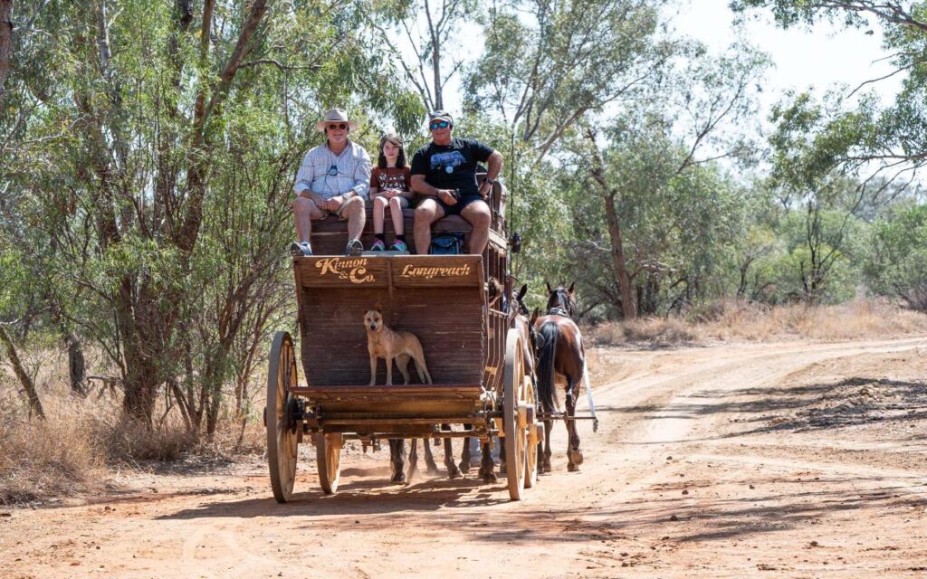Three people sit on a wooden horse-drawn carriage with a dog painted on the front, traveling on a dirt path surrounded by trees.