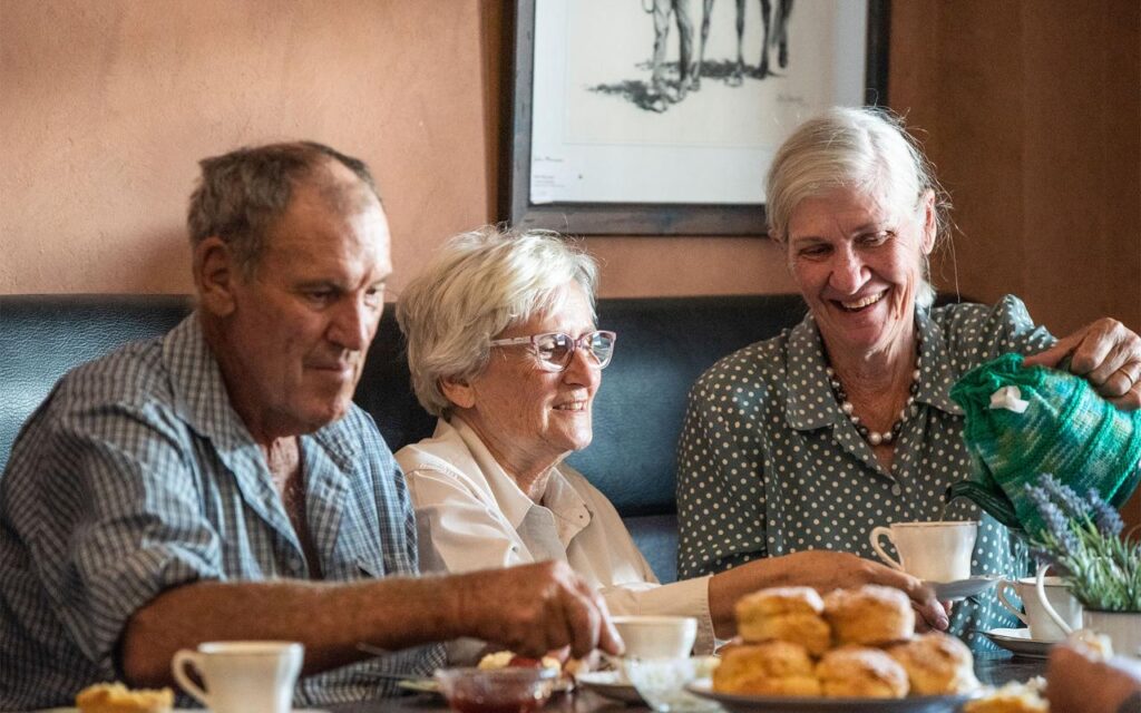 Three older adults sit at a table with tea and scones, smiling and enjoying each other’s company in a warmly lit room.
